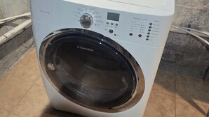 A front-loading washing machine with a control panel, located in a room with tiled flooring and a concrete wall.