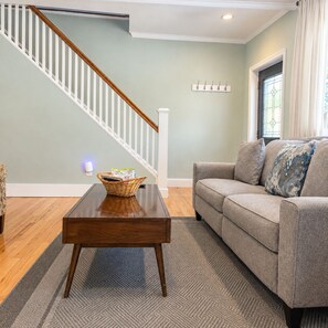 A living room with a wooden coffee table, a grey sofa, a staircase with white railings, and a window with curtains.