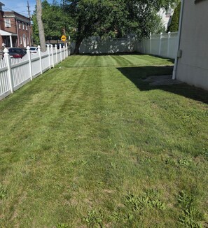 A well-maintained lawn with a white picket fence, a tree, and a building in the background. - Family-friendly 3-bedroom/2.5-Bath house Near NYC and FIFA MetLife Stadium (Teaneck)