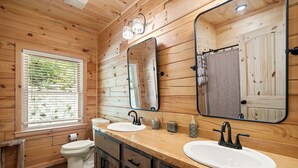 Une salle de bain aux murs lambrissés, équipée de deux lavabos, de toilettes et d’une fenêtre avec des stores.