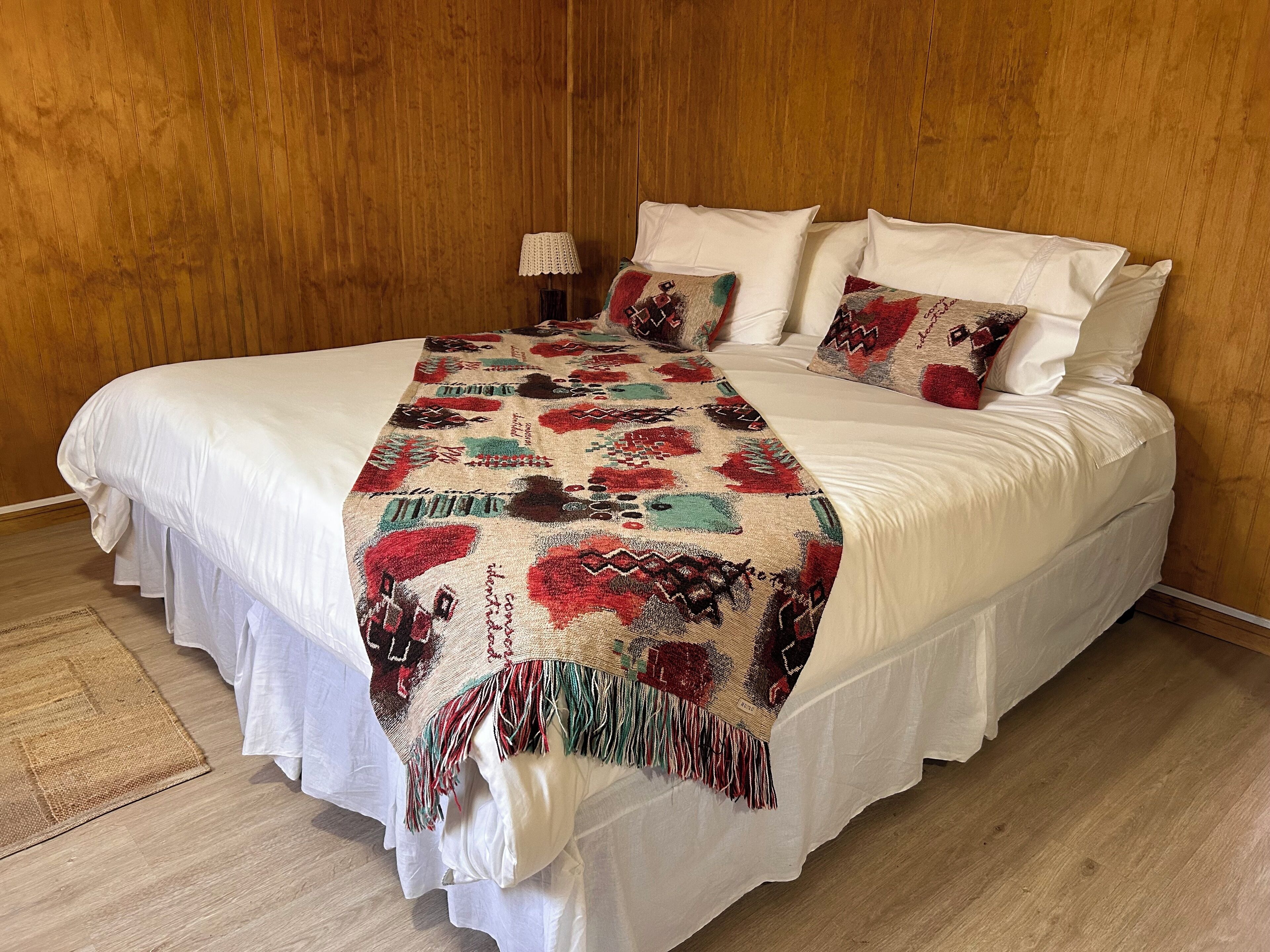 A bed with a white bedspread and a colorful blanket, a small table with a lamp, and a wooden floor.