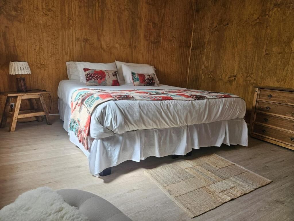 Un dormitorio con pared de madera, una cama con cubrecama y almohadas blancas, una mesita de noche de madera con lámpara y un aparador de madera.