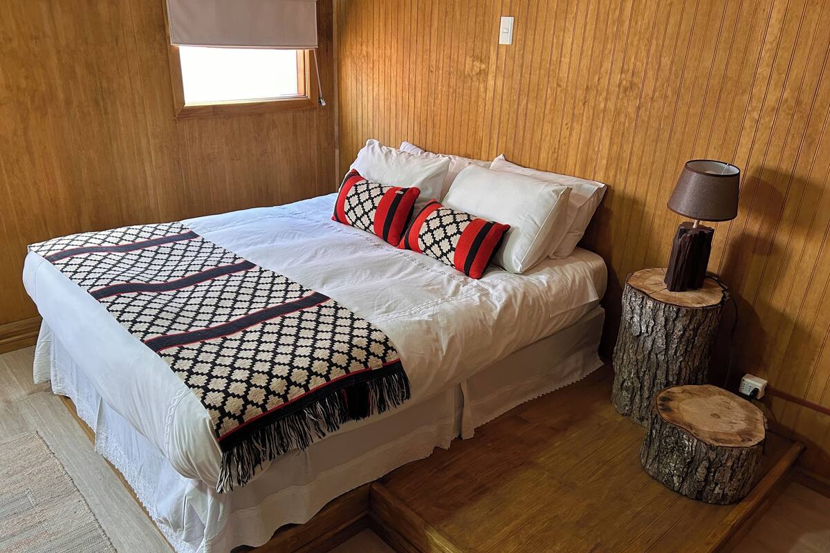 Una cama con una colcha estampada, almohadas blancas y una almohada roja y negra. Hay una lámpara sobre un tocón de árbol junto a la cama.