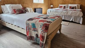 A room with a wooden floor, a bed with a patterned blanket, a second bed with a white bedspread, and a window with a white blind. - Hostal Lago Cóndor Patagonia (Puerto Natales)