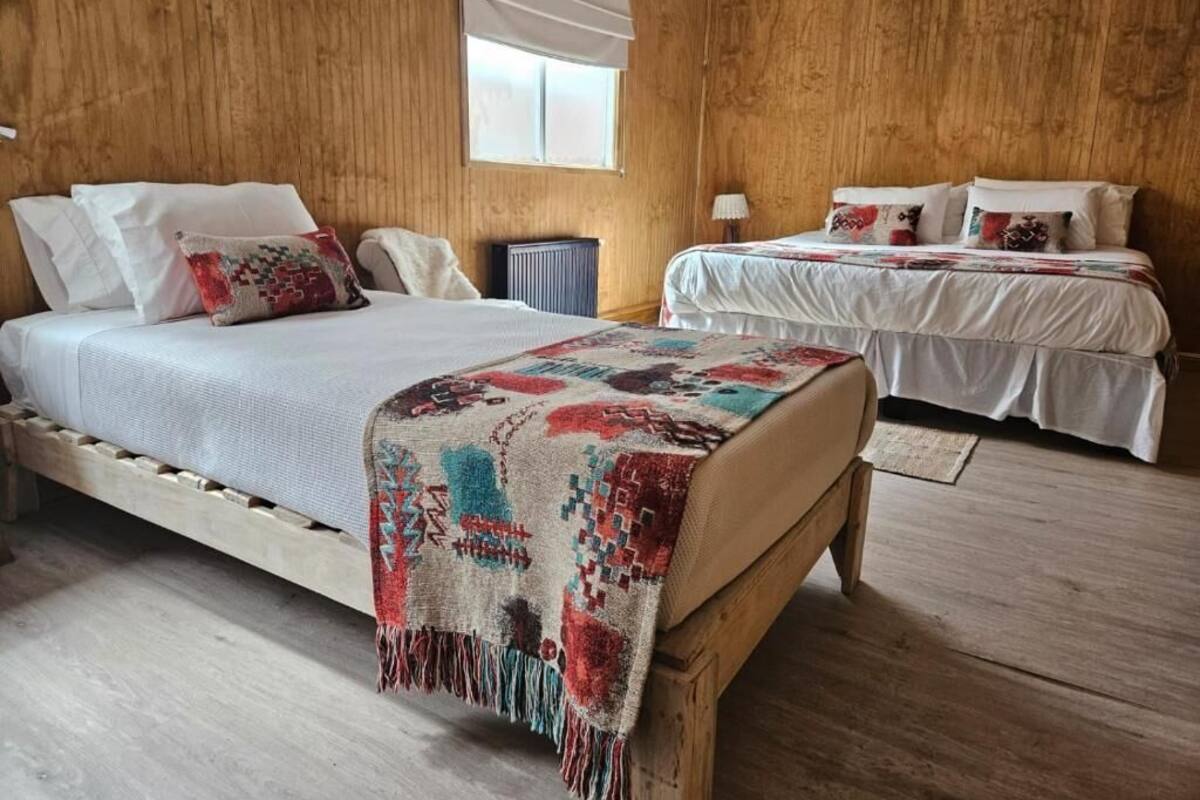 Una habitación con suelo de madera, una cama con una colcha estampada, otra cama con una cubrecama blanca y una ventana con una persiana blanca.