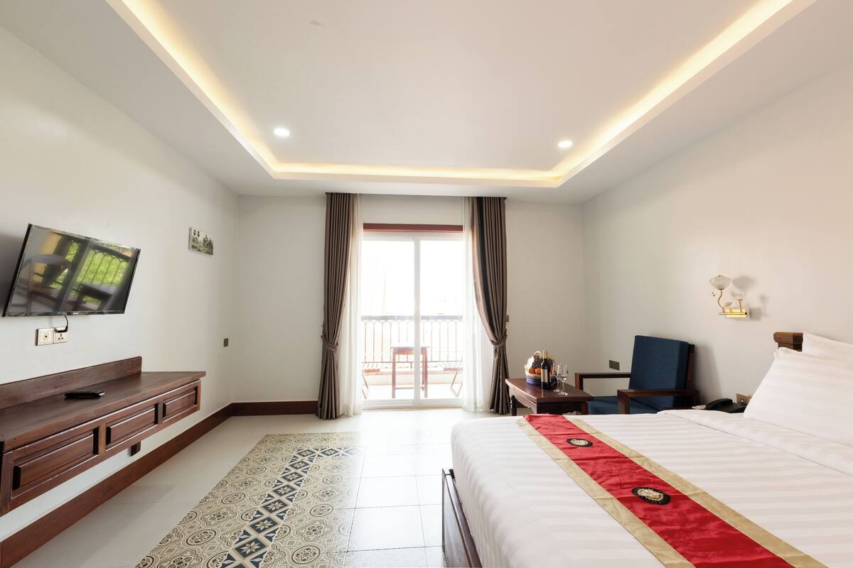 Une chambre d’hôtel comprenant un lit, une télévision, un bureau avec une chaise, une petite table avec une lampe et un balcon avec des rideaux.