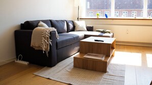 A modern living room with a dark blue sofa, a wooden coffee table, and a large window with a view of buildings. - Stylish Central 4-person CPH Apt. (København)