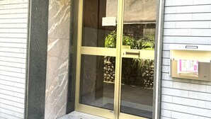 Una entrada de edificio con puerta de cristal, buzón y un pequeño espacio exterior con muro de piedra y plantas.