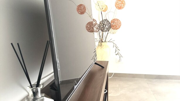 Una habitación moderna con una televisión de pantalla plana sobre un soporte de madera, una botella de vidrio con la etiqueta 'LUXURY' en el soporte, y un florero con elementos decorativos esféricos sobre una mesa.