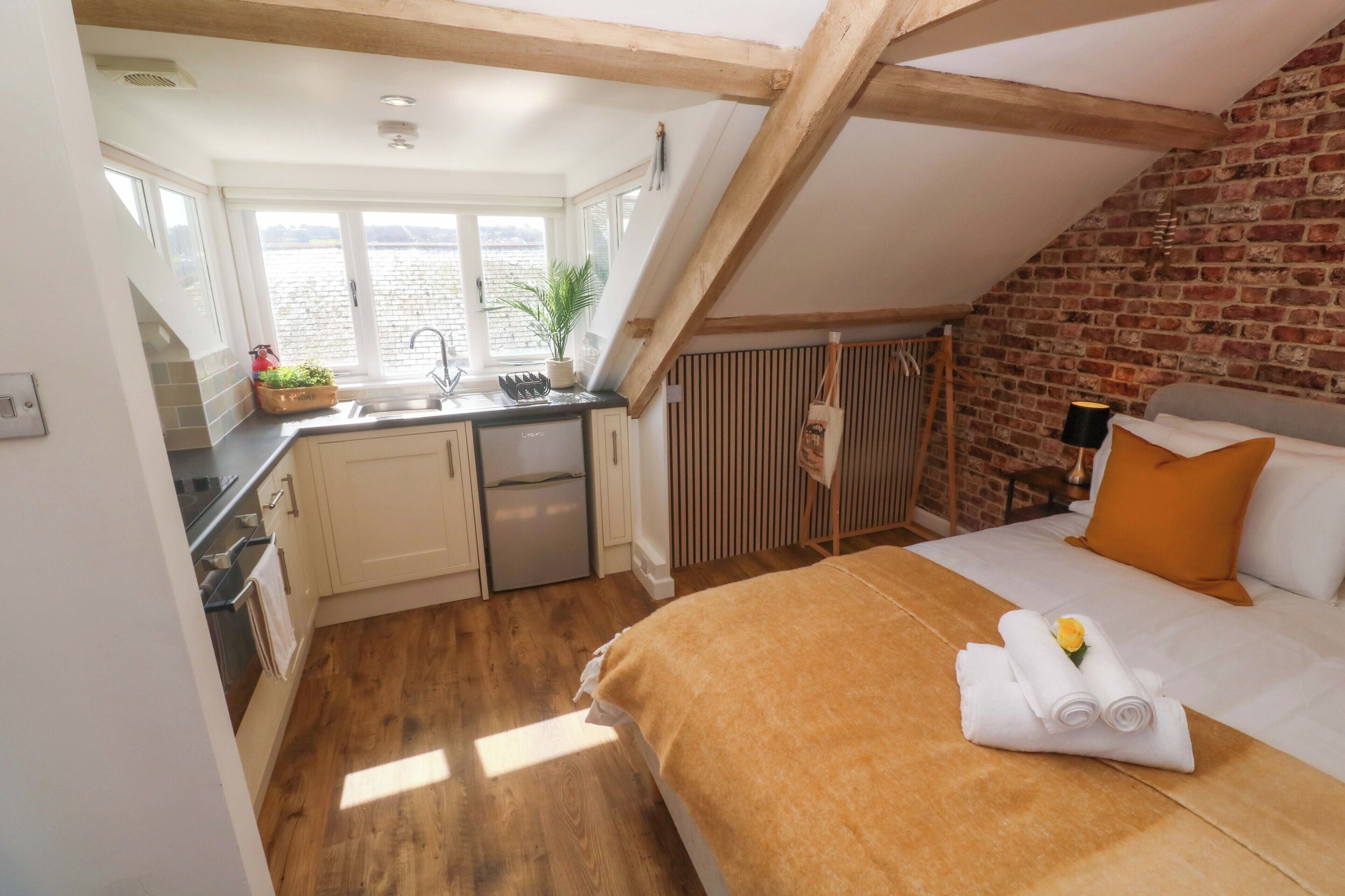 A bedroom with a bed, a window, a kitchen area, and a brick wall.