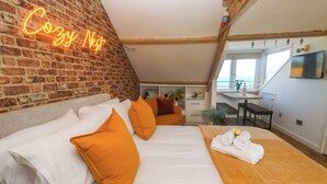A cozy bedroom with a brick wall, a bed with white and orange bedding, a small desk with a chair, and a TV mounted on the wall. - Seagulls Nest (St Ives)