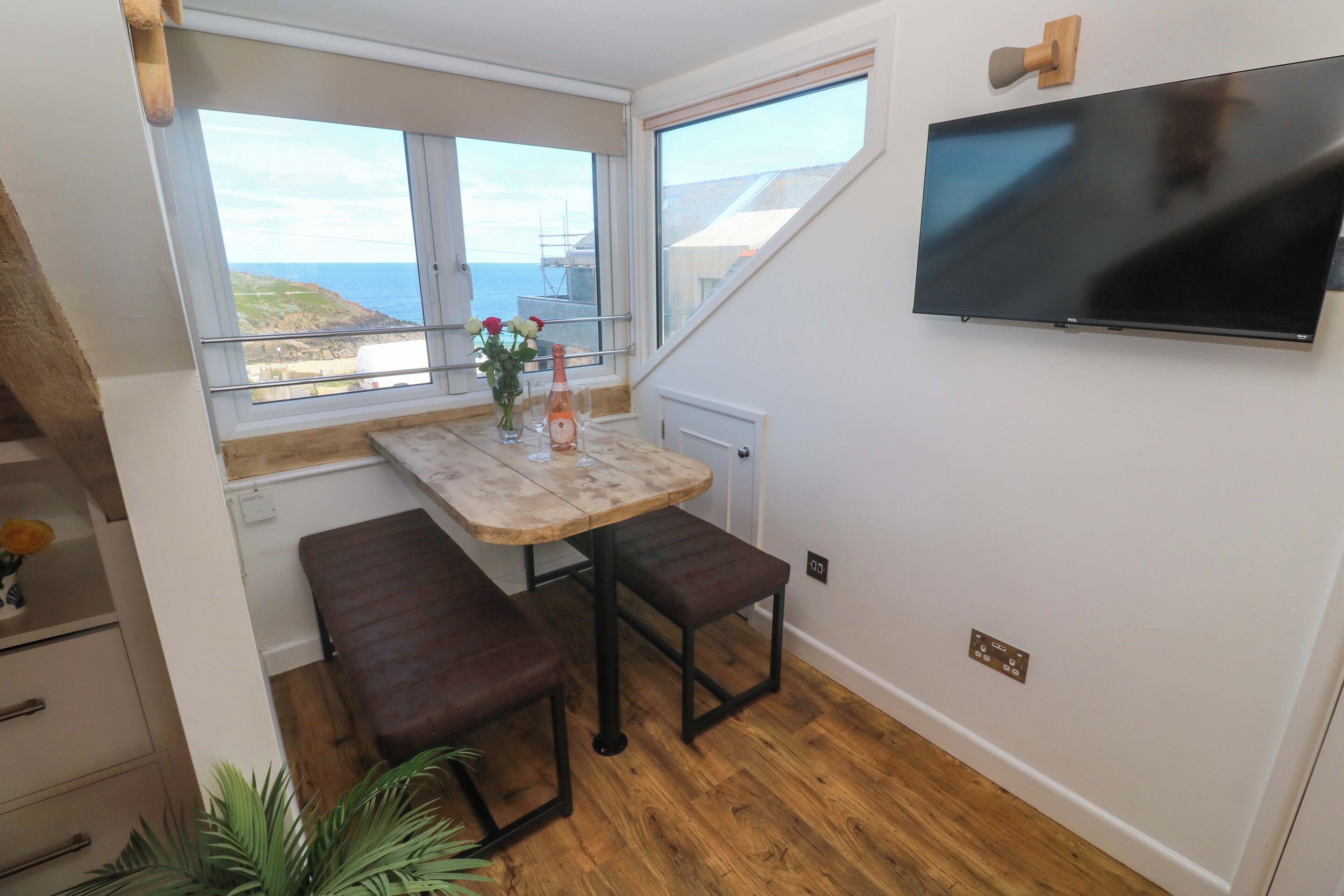 A small room with a wooden table, bench seating, a TV mounted on the wall, and a window with a view of the sea.