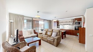 Ein modernes Wohnzimmer mit Sofa, Couchtisch und Korbstühlen. Im Hintergrund ist eine Küche mit Mikrowelle und Herd erkennbar.