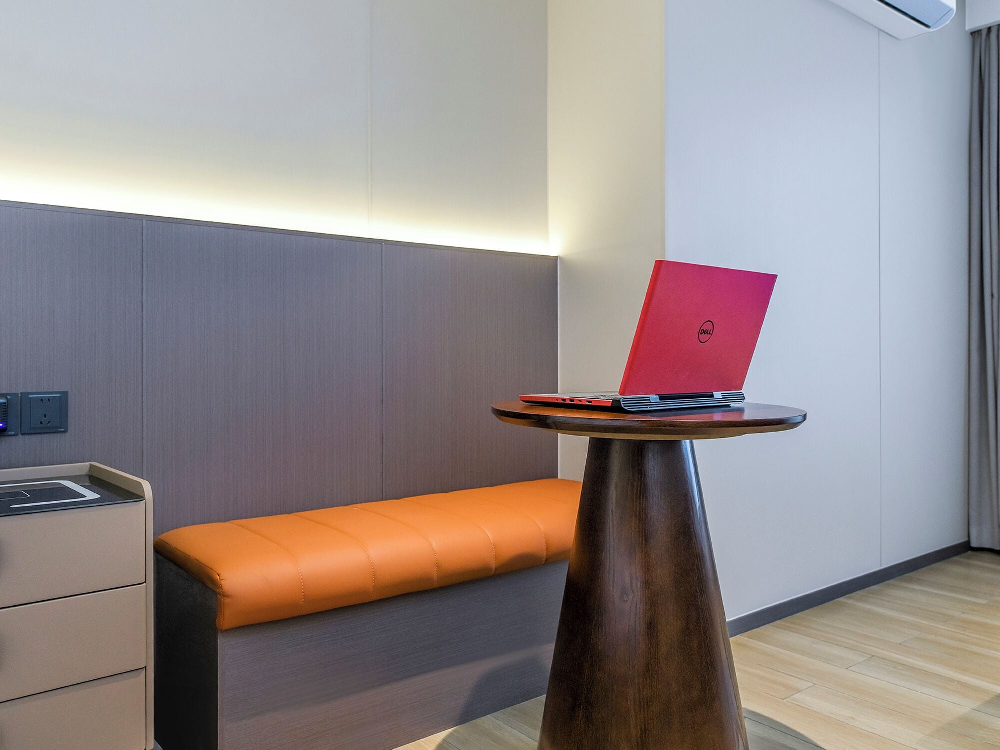 Ein modernes Hotelzimmer mit Holzboden, einer grauen gepolsterten Bank, einem kleinen runden Tisch mit einem roten Laptop und einer Wand-Klimaanlage.