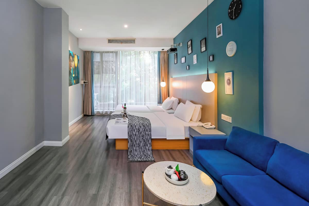 Una habitación de hotel moderna con una cama, un sofá azul, una mesita redonda con un objeto decorativo y un ventanal con cortinas translúcidas.
