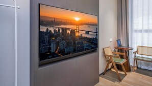 Habitación de hotel moderna con una televisión grande que muestra un paisaje urbano al atardecer, una silla de madera, una mesita con una computadora portátil y una ventana con cortinas.