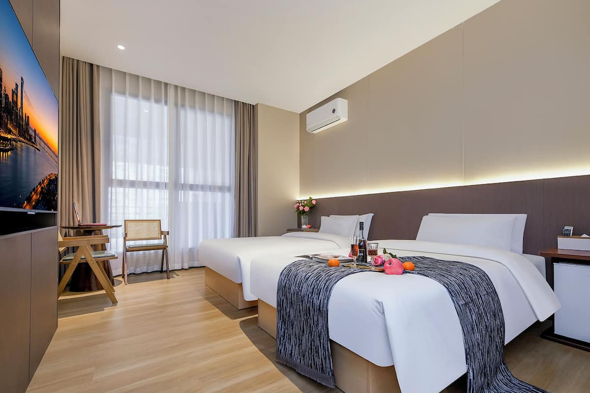 Habitación de hotel con dos camas, un escritorio, una silla, un televisor de pantalla plana y un ventanal con cortinas.