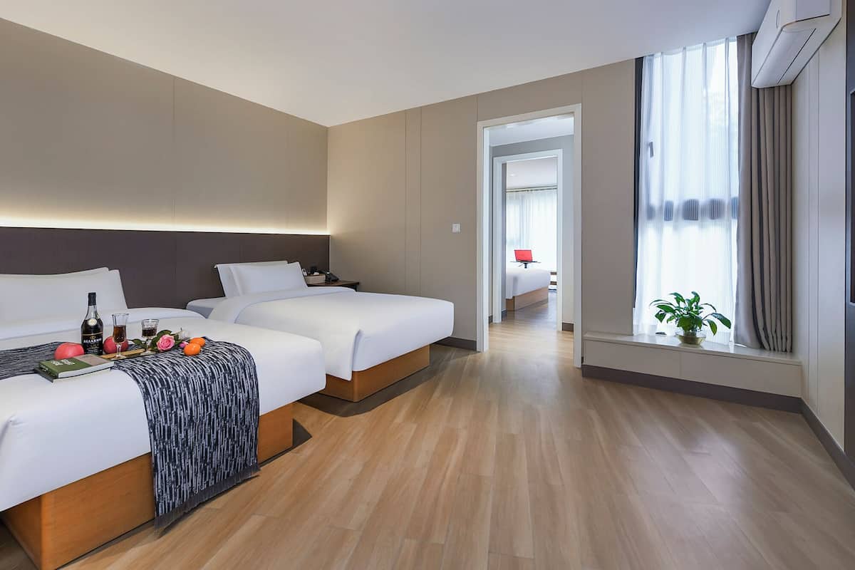 Habitación de hotel con dos camas, una mesita de noche con una botella y vasos, una planta en maceta junto a la ventana y vistas a otra habitación a través de una puerta abierta.