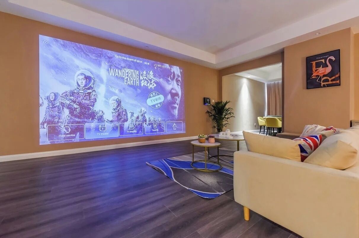 A hotel lobby with a large digital screen displaying a movie poster, a sofa, a small table with plants, and a framed artwork on the wall.