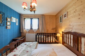 A bedroom with a wooden headboard, a bed with a floral bedspread, a desk with a chair, a radiator, and a window with curtains.
