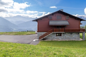 A red wooden house with a stone foundation, a wooden balcony, and a paved driveway.