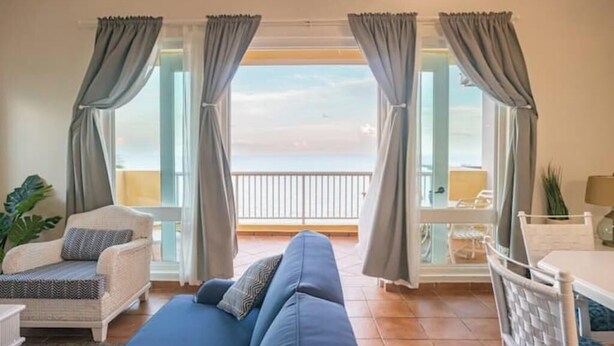 Ein Wohnzimmer mit einer Couch, einem Sessel, einem Tisch und Meerblick durch große Fenster.