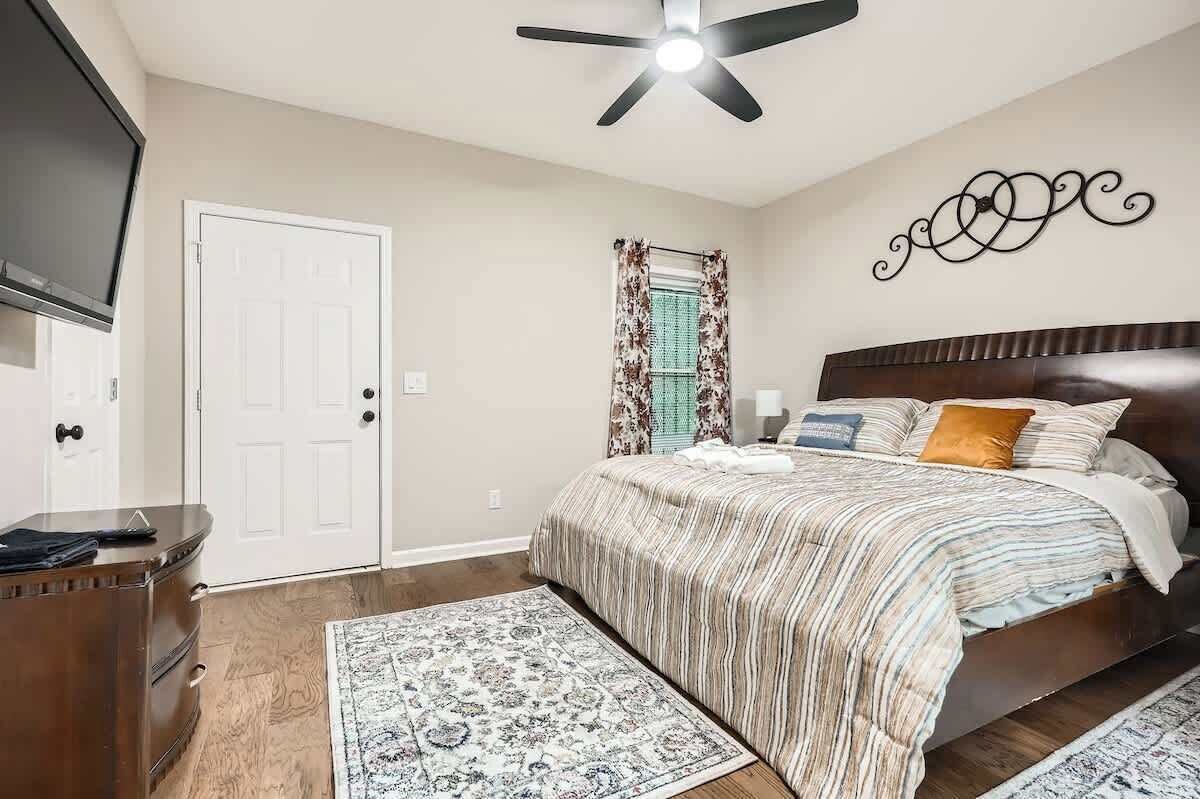 A bedroom with a bed, a ceiling fan, a TV, a dresser, a rug, and a window with curtains.