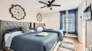 Une chambre avec deux lits, un ventilateur de plafond, des œuvres d’art décoratives au mur et une grande fenêtre avec des rideaux.