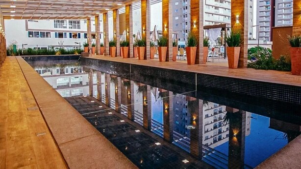 A long indoor swimming pool with a glass ceiling, potted plants, and a view of surrounding buildings.