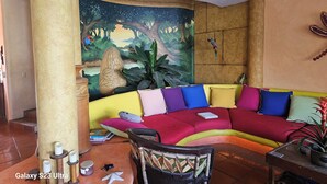 Un salon accueillant avec un canapé coloré, un ventilateur de plafond, une fresque murale et une petite table sur laquelle est posée une plante.