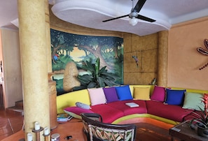 Uma sala de estar acolhedora com um sofá colorido, um ventilador de teto, um mural e uma pequena mesa com uma planta.