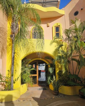 Um edifício amarelo com entrada em arco, rodeado por plantas em vasos e palmeiras.
