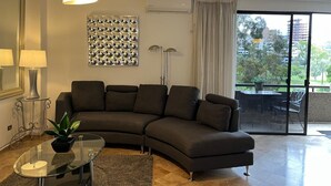 A modern living room with a curved sofa, a glass coffee table, and a large window with a view of greenery and buildings. - Charming, elegant cozy 1-bedroom condo in amazing Tijuana with AC main Blvd SAFE (Tijuana)