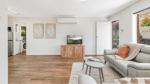 Ein modernes Wohnzimmer mit einer Couch, einem Couchtisch und einem Fernseher auf einem Holzboden. Im Hintergrund ist eine Küchenzeile mit einer Waschmaschine zu sehen.