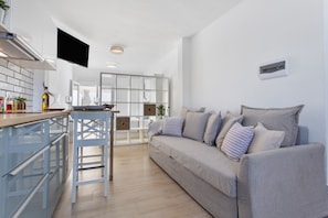 A modern kitchen with a flat-screen TV, a bar area, a grey sofa, and a dining area with a white table and chairs.