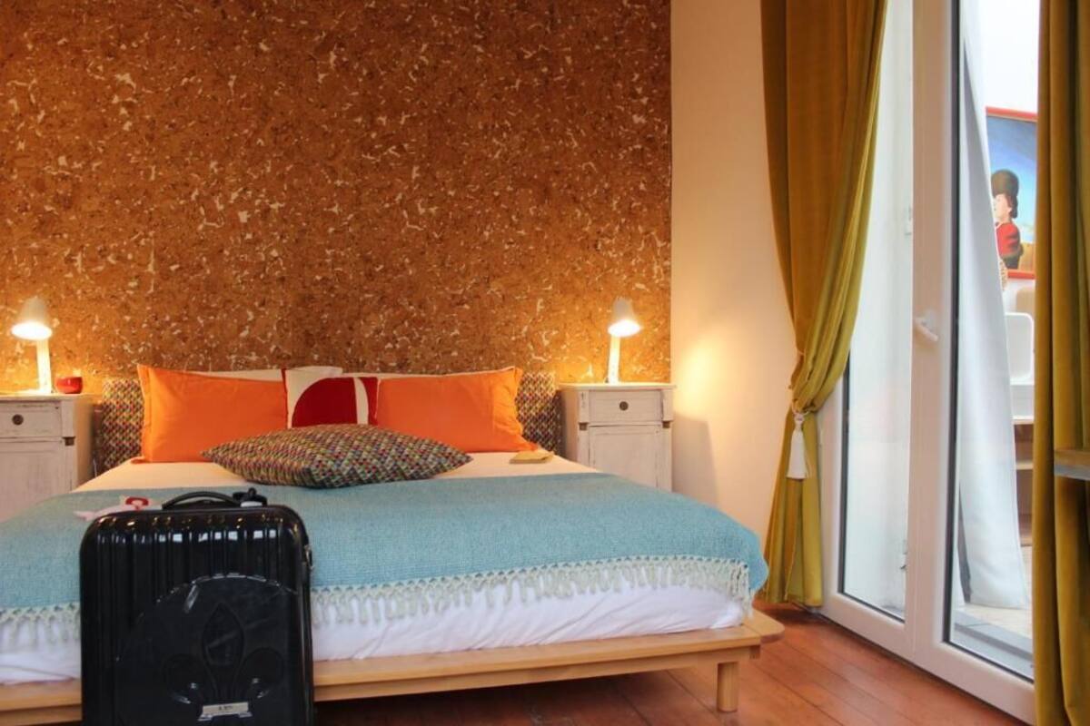 Un lit bien fait, avec une valise posée dessus, deux tables de chevet, une fenêtre avec des rideaux et un mur à texture.