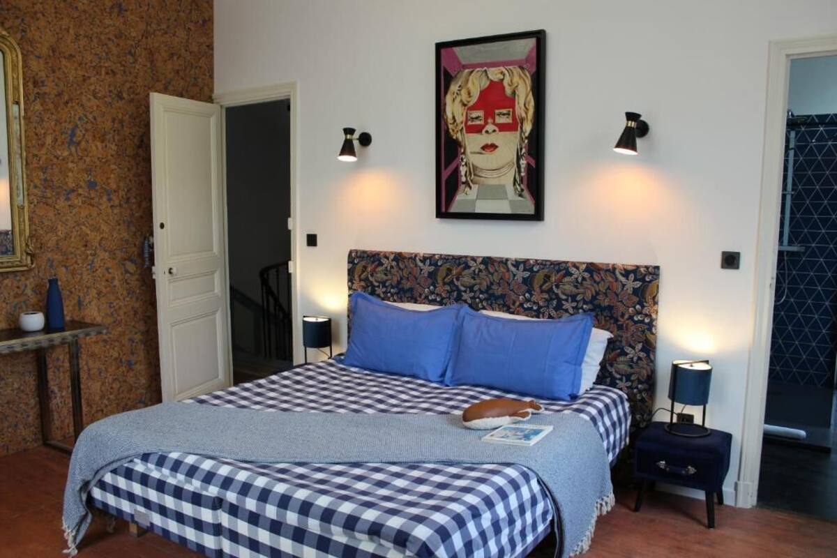 Une chambre à coucher avec une couvre-lit à carreaux, une tête de lit à motifs floraux, deux lampes fixées au mur et une œuvre d’art encadrée au-dessus du lit.