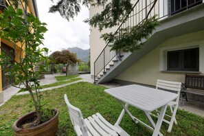 Une cour avec une table et des chaises blanches, un pot de fleurs et un escalier menant à l’étage supérieur.