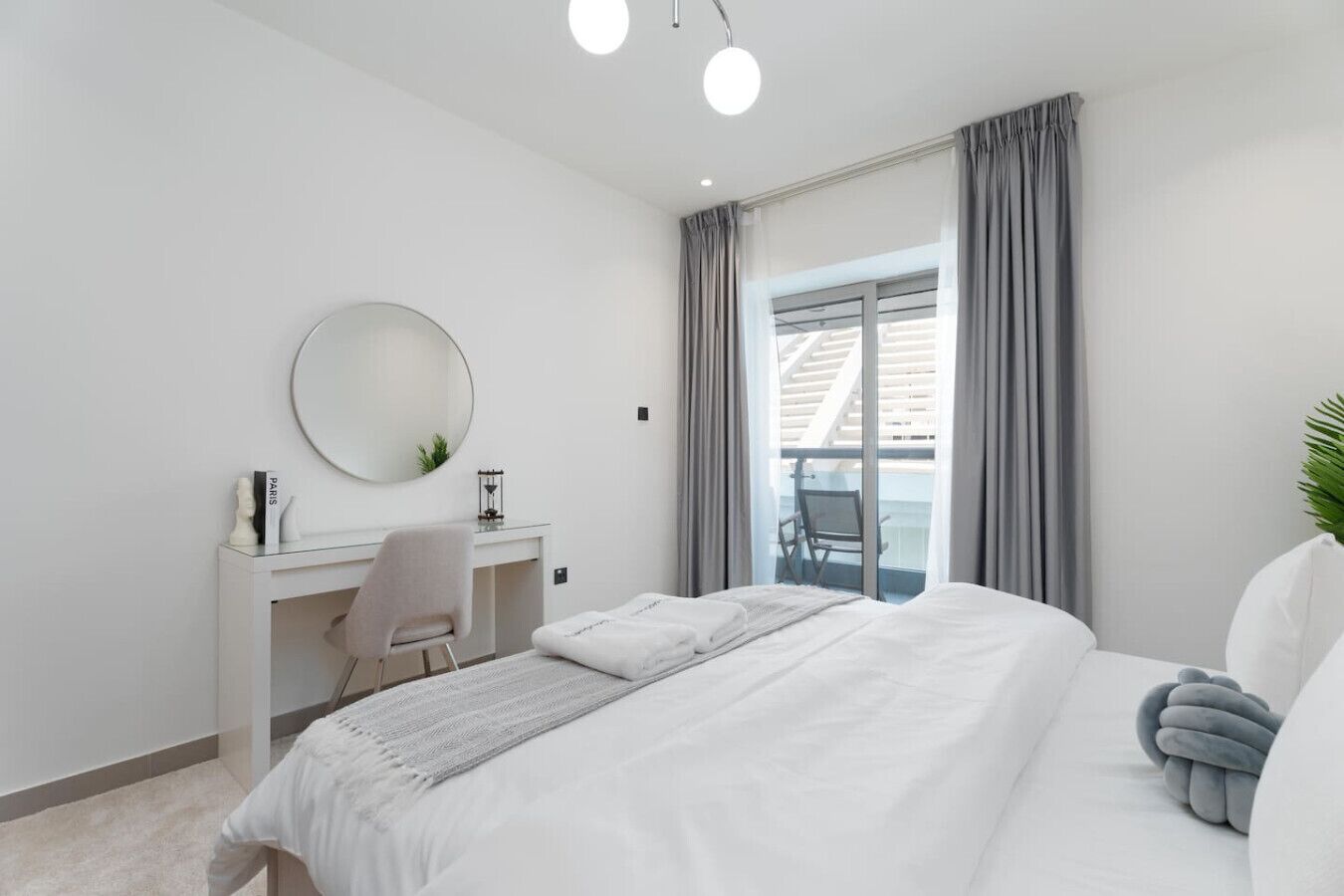 Ein modernes Hotelzimmer mit einem großen Bett, einem Schreibtisch mit Stuhl, einem runden Spiegel und Blick ins Freie durch ein Fenster mit grauen Vorhängen.