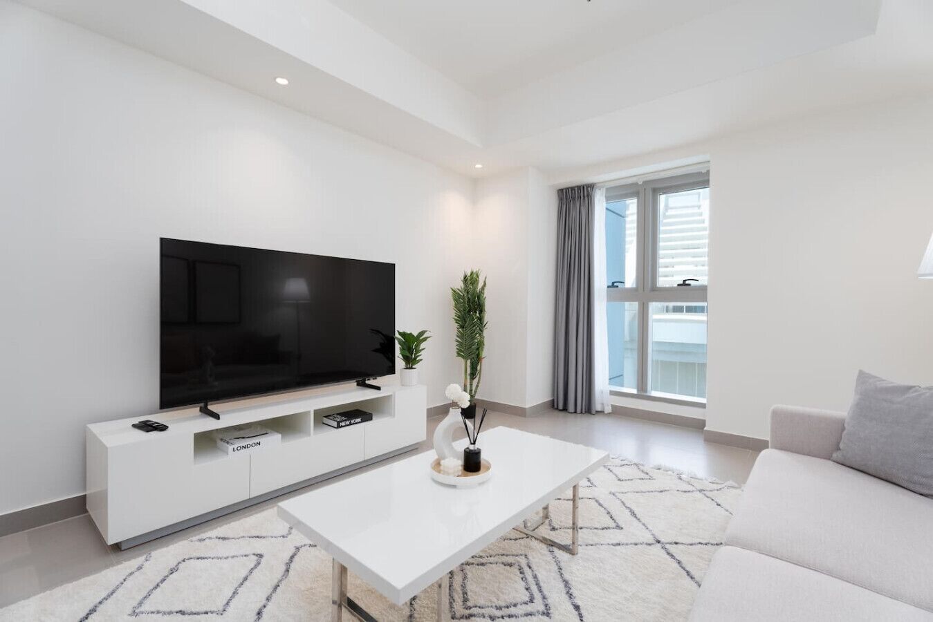 A modern living room with a flat-screen TV, a white coffee table, a sofa, and a large window with curtains.