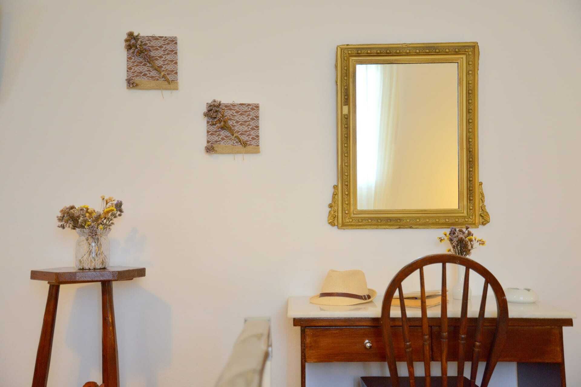 Een kamer met een houten stoel, een tafeltje met een bloevas en een bureau met een hoed erop. Er hangen drie ingelijste foto's aan de muur.