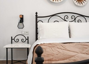 Un lit bien fait, avec du linge de lit blanc et une couverture marron, une petite table de chevet avec une lampe, et une prise de courant fixée au mur.