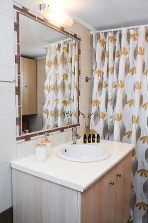 Une salle de bain avec un lavabo blanc, des meubles en bois, un miroir et un rideau de douche orné d’un motif de feuilles.