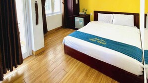 Una camera d'albergo con pavimento in legno, un letto con biancheria azzurro e bianca, una parete gialla a contrasto, una ventola a soffitto e una finestra con tende.