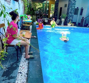 A swimming pool with people in the water and on the edge, surrounded by plants and a covered seating area. - VT Villa Cua Dai Hoi An (Hoi An)