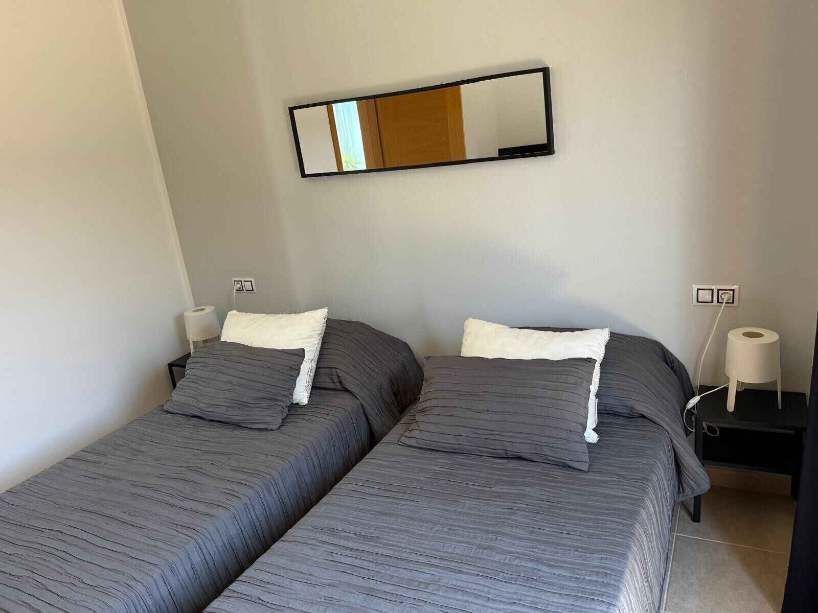 A room with two beds, each with a pillow and a bedside table with a lamp.