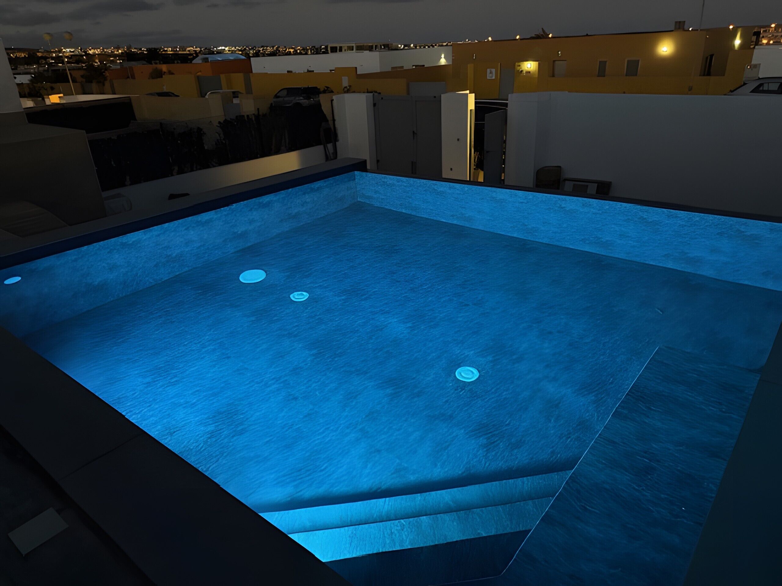 A rooftop swimming pool with a blue-lit interior, surrounded by buildings.