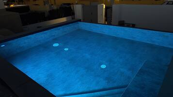 A rooftop swimming pool with a blue-lit interior, surrounded by buildings.