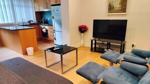 A modern living room with a sofa, a TV, a small table, and a kitchen area in the background.