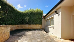 A paved courtyard with a brick wall, a hedge, and a building with a window and a wall-mounted light fixture.